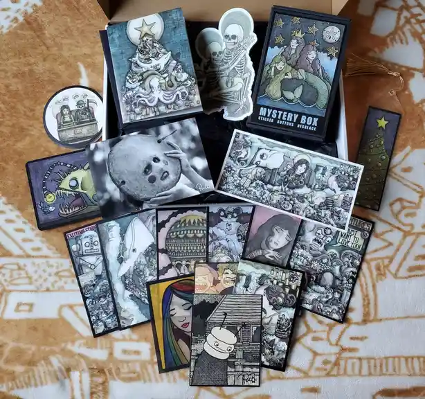 An open cardboard box spills out numerous intricately illustrated cards and stickers featuring surreal, darkly whimsical art…