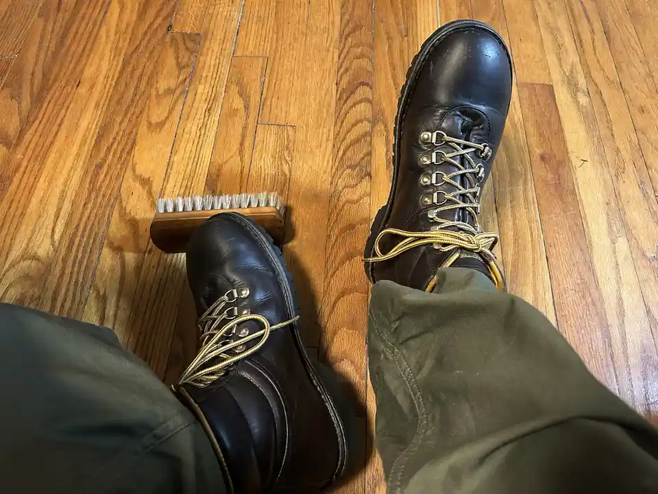A first-person view shows a person's feet in dark brown lace-up boots resting on a wooden floor, with a wooden shoe brush…