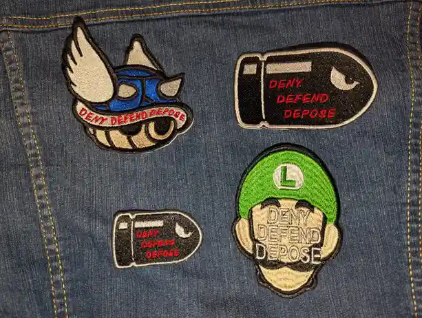 Four embroidered patches are sewn onto blue denim jeans. One patch shows a cartoon character with wings and a mask, another…