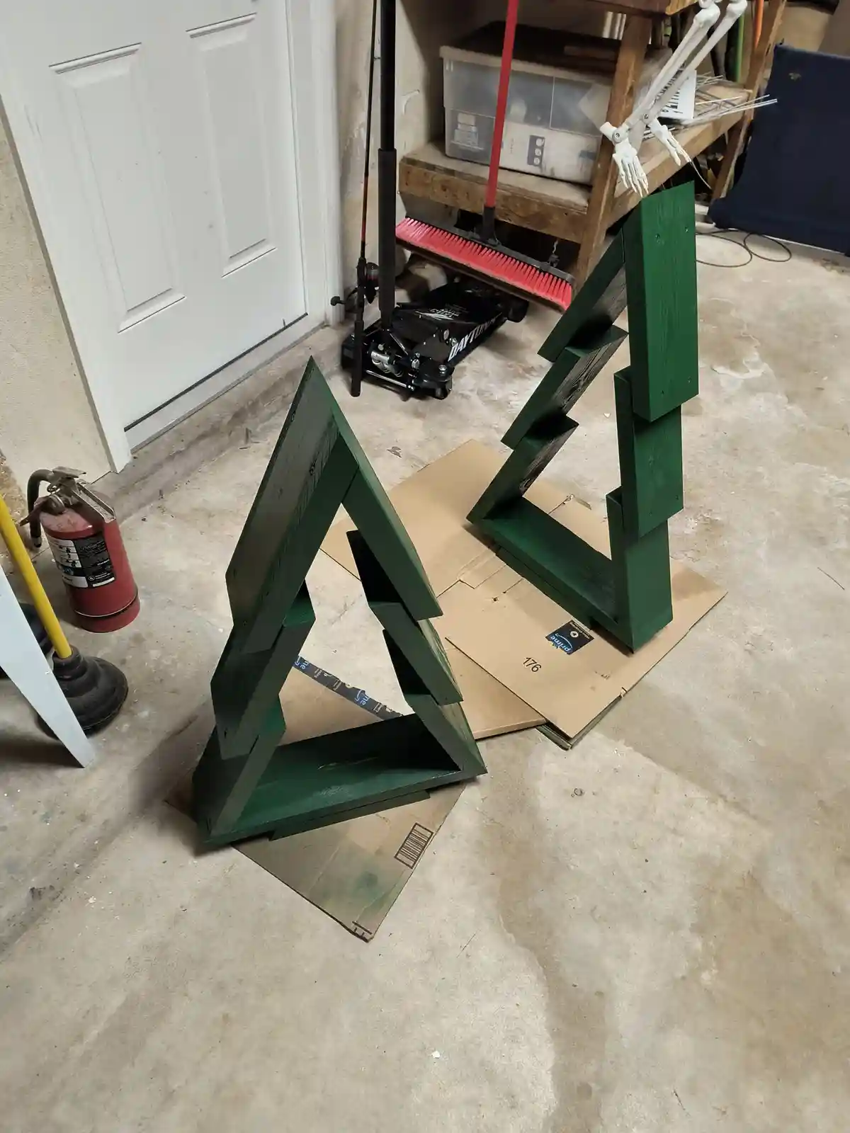 Two green-painted wooden structures shaped like Christmas trees stand on cardboard boxes in a cluttered garage, surrounded…