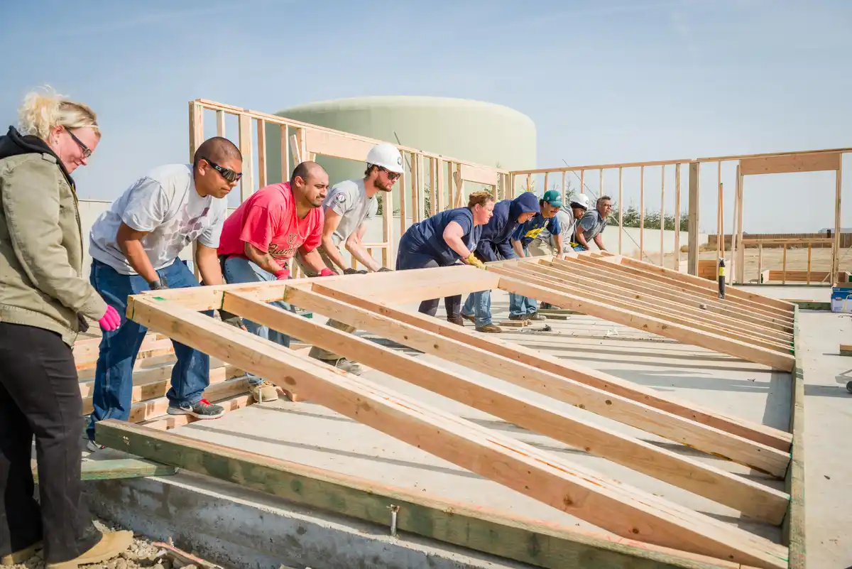 A group of people, including a woman in a green jacket and pink gloves, are pushing heavy wooden beams on a construction…