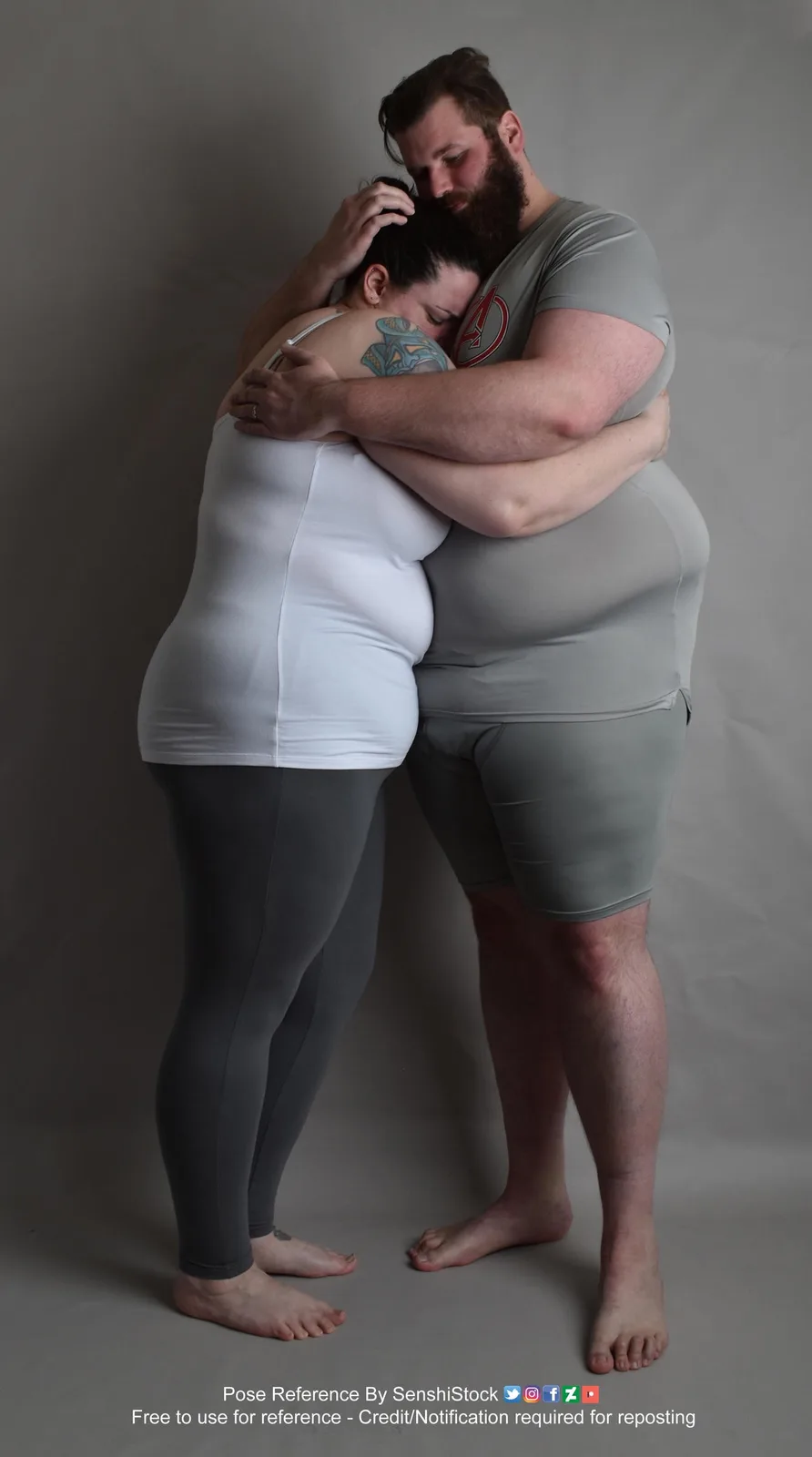 A man and a woman are embracing in a studio setting against a plain gray background. The man, wearing a gray t-shirt with an…