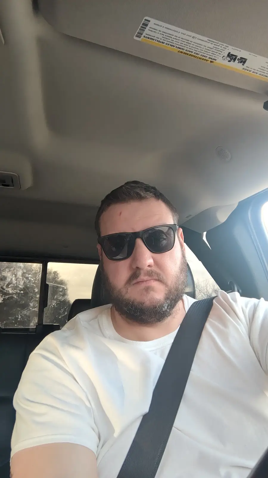 A man with a beard and sunglasses sits in the driver's seat of a car, wearing a white t-shirt and seatbelt, looking directly…