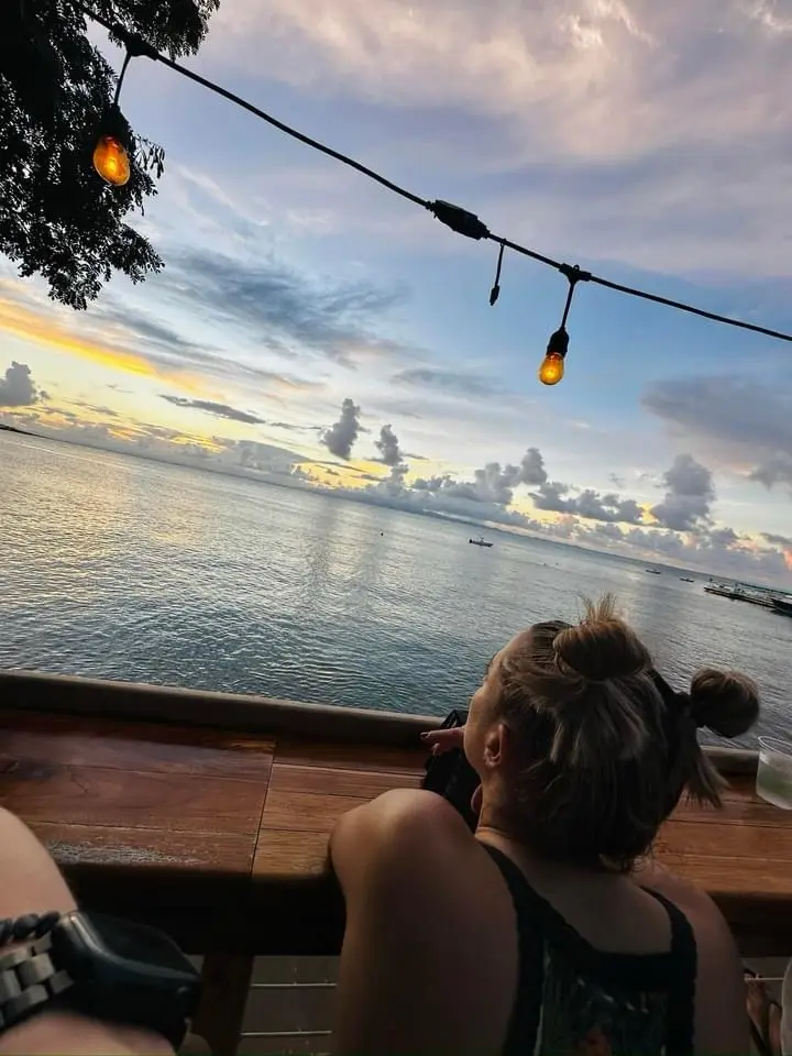 A person with their hair in two buns sits on a wooden railing overlooking a calm ocean at sunset, with string lights hanging…