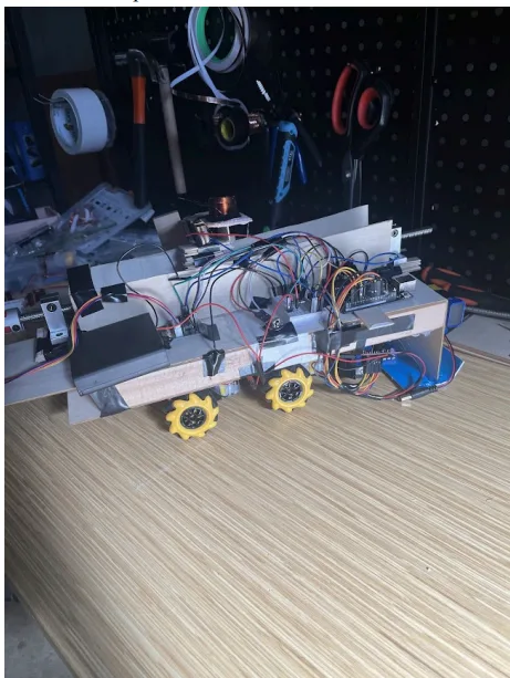 a cluttered, homemade robot with exposed wires, circuit boards, and yellow wheels sits on a wooden surface, surrounded by…