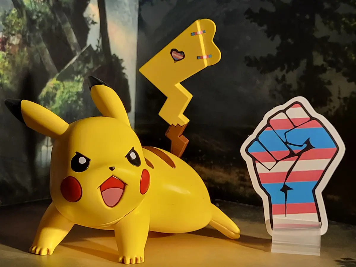 A yellow Pikachu figurine with an angry expression is shown next to a raised fist and a heart-shaped sticker, both decorated…