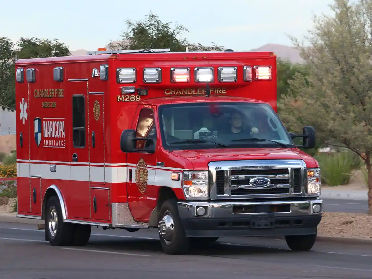A red Chandler Fire / Maricopa Ambulance, marked M289, is shown driving on a road with trees and hills in the background…