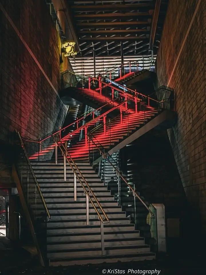 A dimly lit, multi-level staircase with red neon lighting on upper steps and brick walls, featuring a yellow sign that reads…