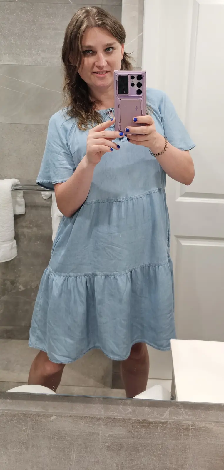 A woman with brown hair and blue nail polish takes a mirror selfie in a bathroom wearing a light blue tiered dress. She…