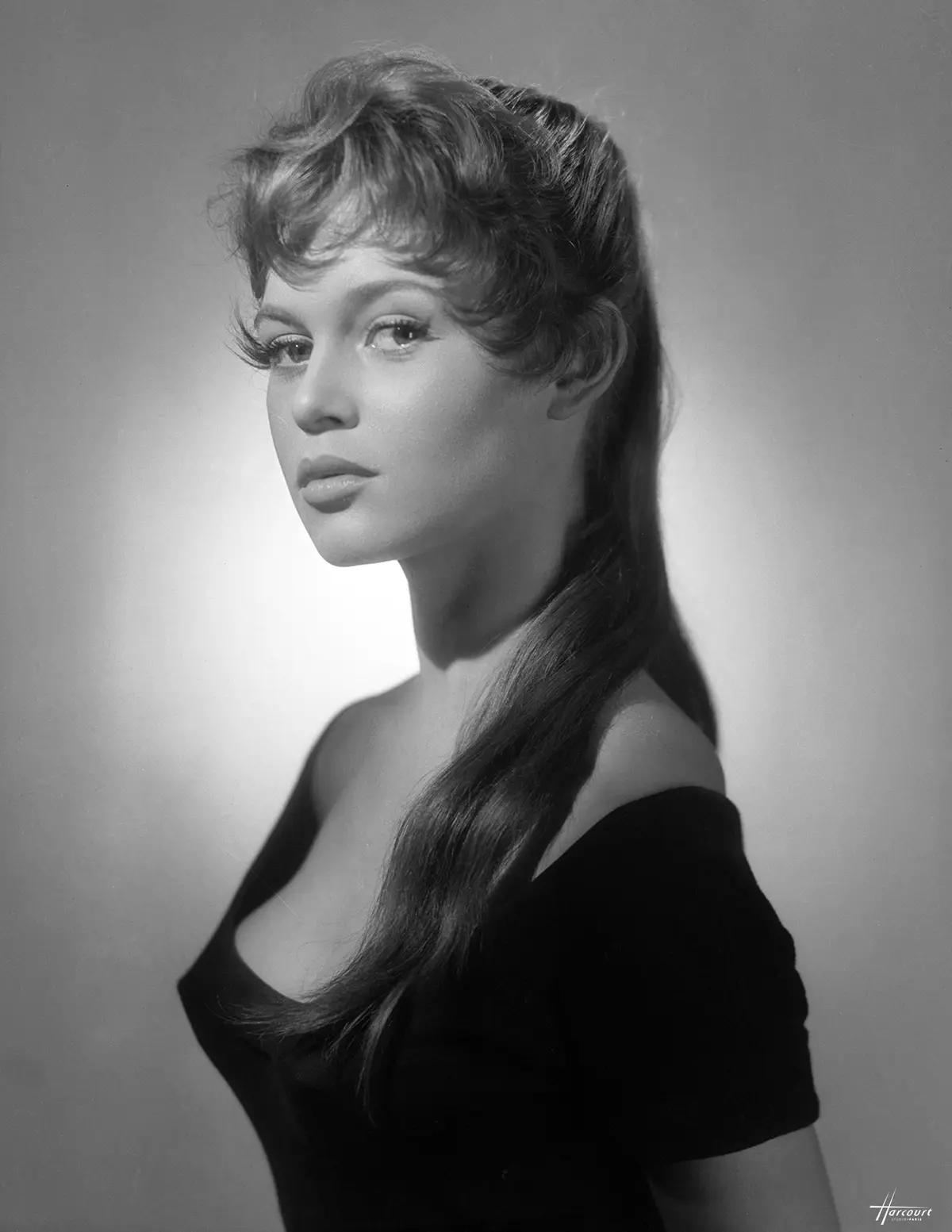 A black-and-white studio portrait of a young woman with long hair and a black off-the-shoulder top, looking over her…
