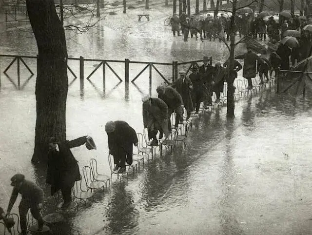 A black-and-white historical photo shows a long line of people wading through floodwaters, each balancing on a metal chair…