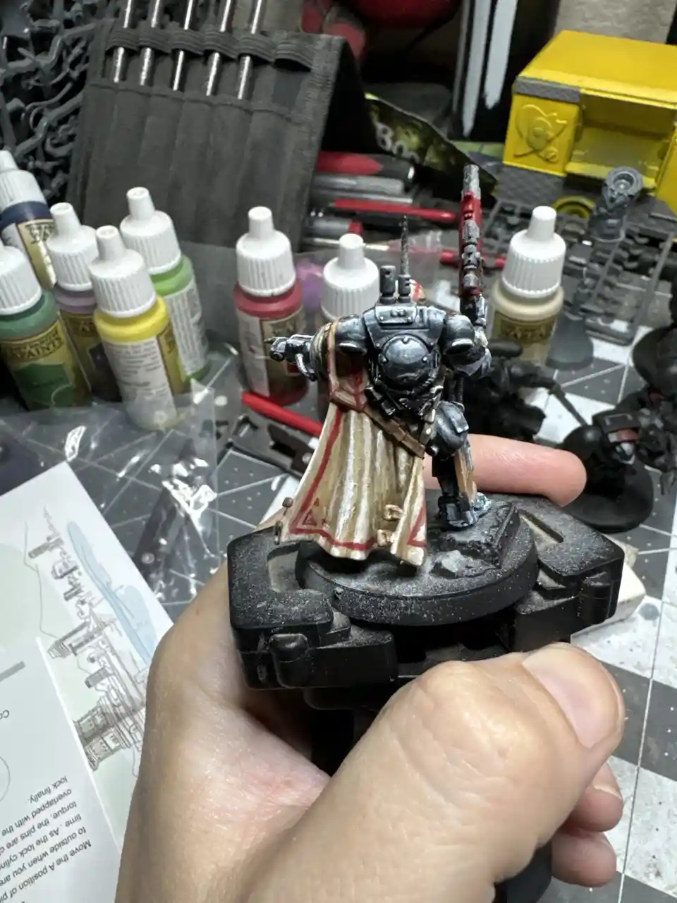 A hand holds a painted miniature of a heavily armored warrior with a flowing cape, surrounded by paint bottles and hobby…