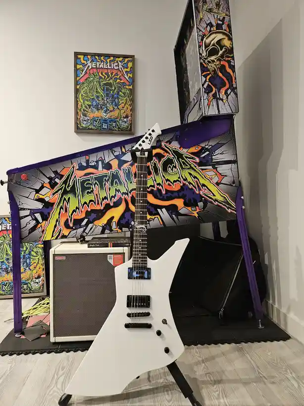 A white electric guitar is propped up in front of a custom-painted Metallica pinball machine, with a framed Metallica poster…