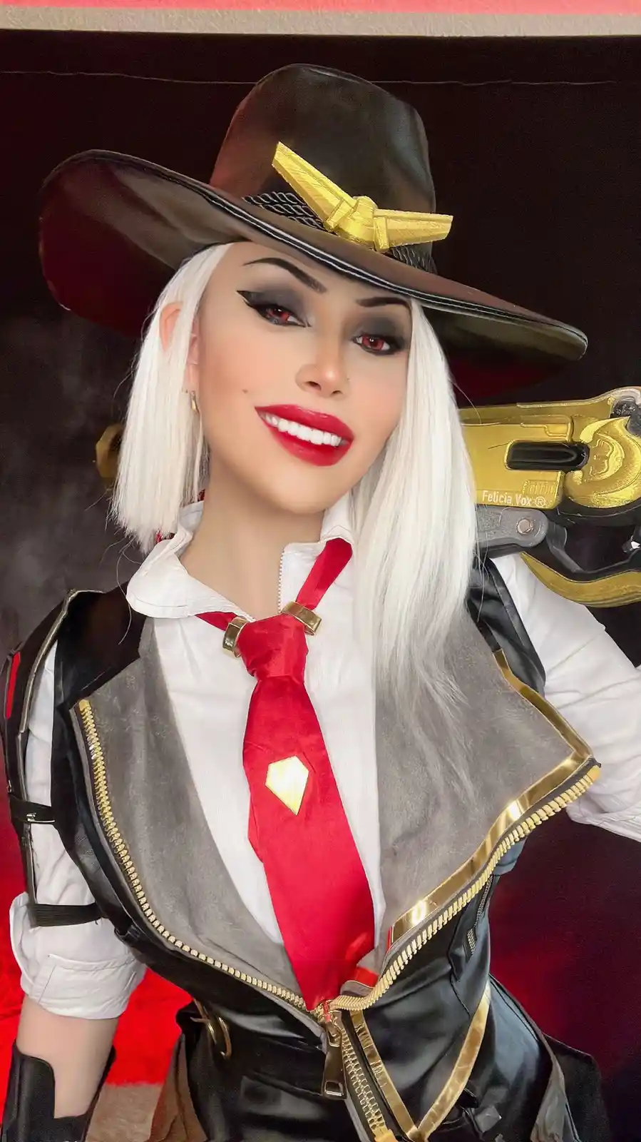 A person in cosplay as the Overwatch character Ashe, wearing a brown cowboy hat with a gold bow, white wig, red lipstick…