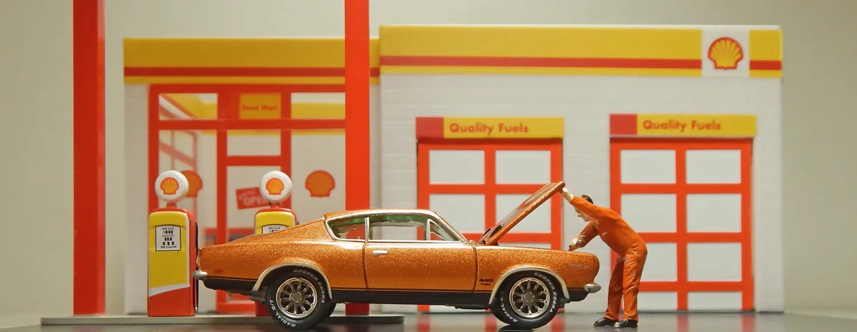A miniature diorama shows a tiny figure in an orange jumpsuit lifting the hood of a glittery orange muscle car at a Shell…
