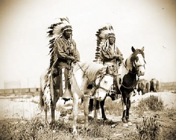 A sepia-toned historical photograph shows two men wearing traditional Native American feathered headdresses, each mounted on…