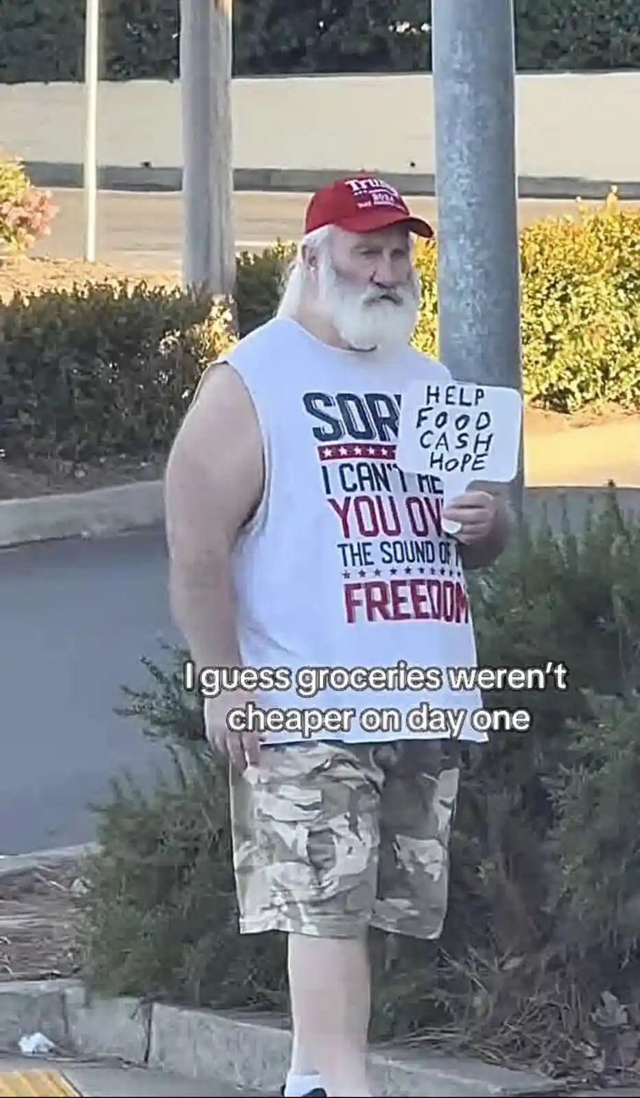 A man with a long white beard and a red 'Trump 2024' cap stands outdoors holding a sign that reads 'HELP FOOD CASH HOPE'. He…