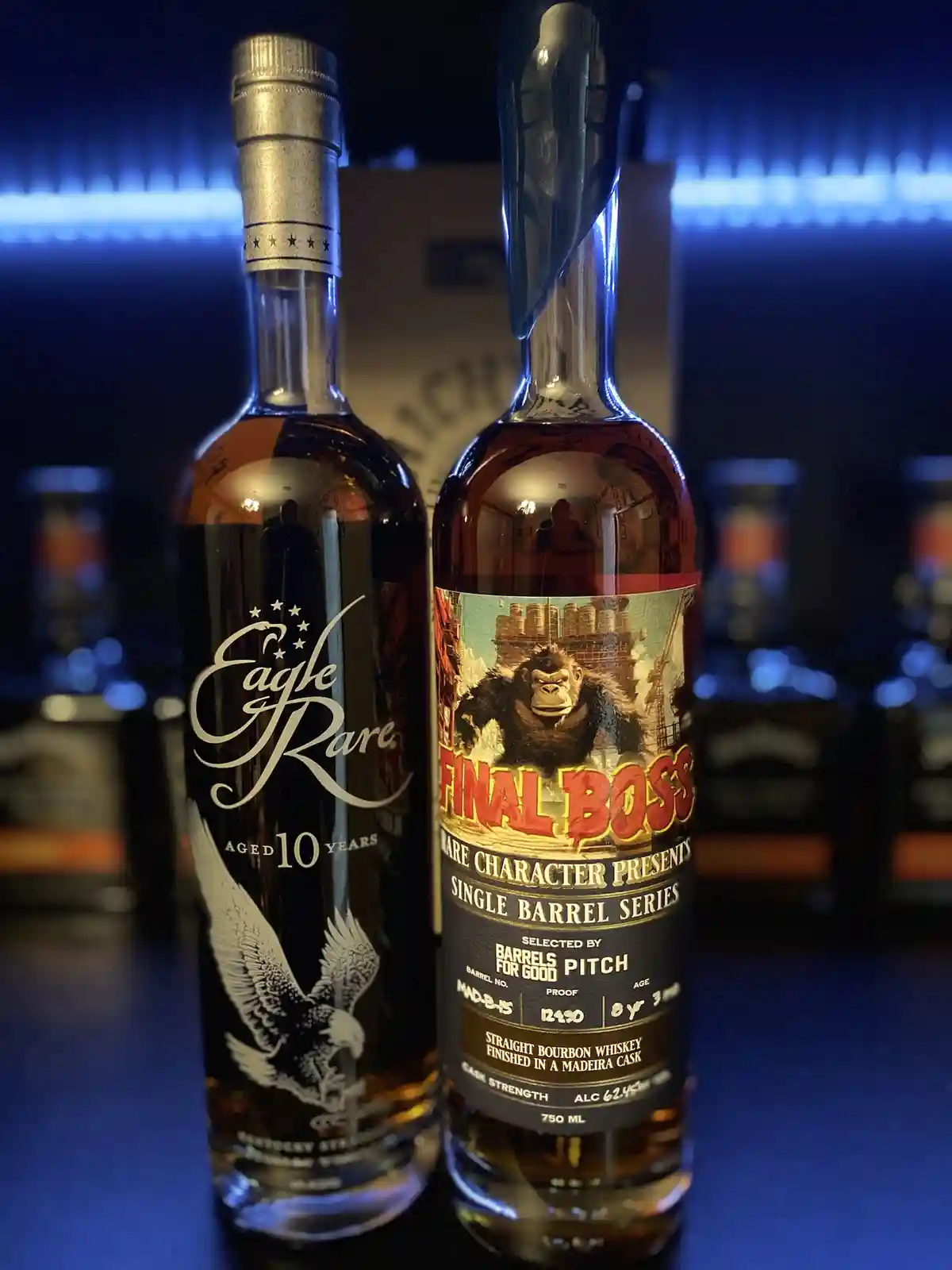 Two whiskey bottles stand side by side on a dark surface under blue lighting; the left bottle is Eagle Rare 10 Years with an…