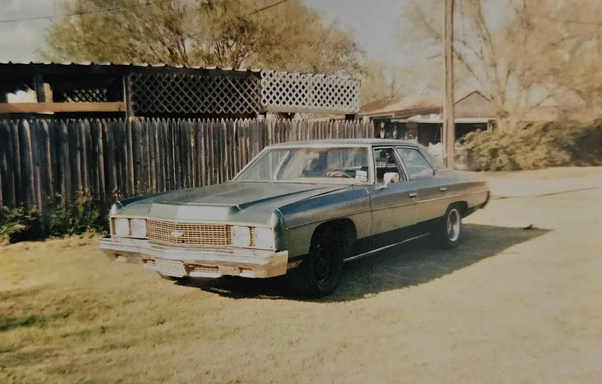 A vintage, slightly rusty blue sedan is parked on a grassy area next to a wooden fence and lattice structure. The car…