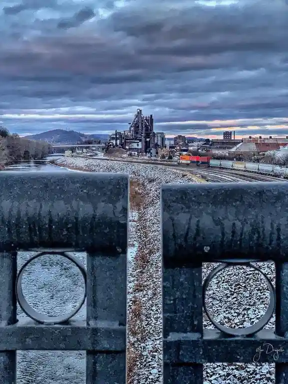 A photograph shows a snowy industrial landscape viewed through the circular openings of a dark metal fence in the…