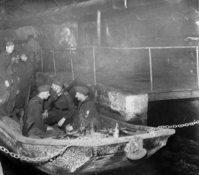 A black-and-white historical photo shows several soldiers in uniform sitting in a small, weathered boat inside a large…