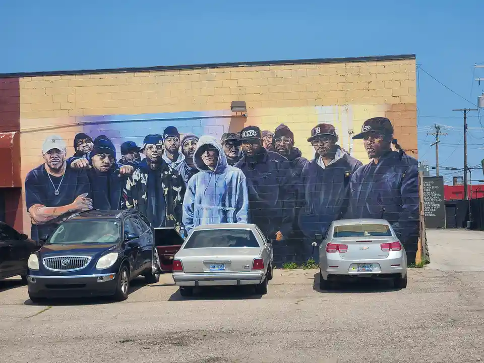 A large mural on a yellow brick wall depicts a group of men, including one in a light blue hoodie standing prominently in…