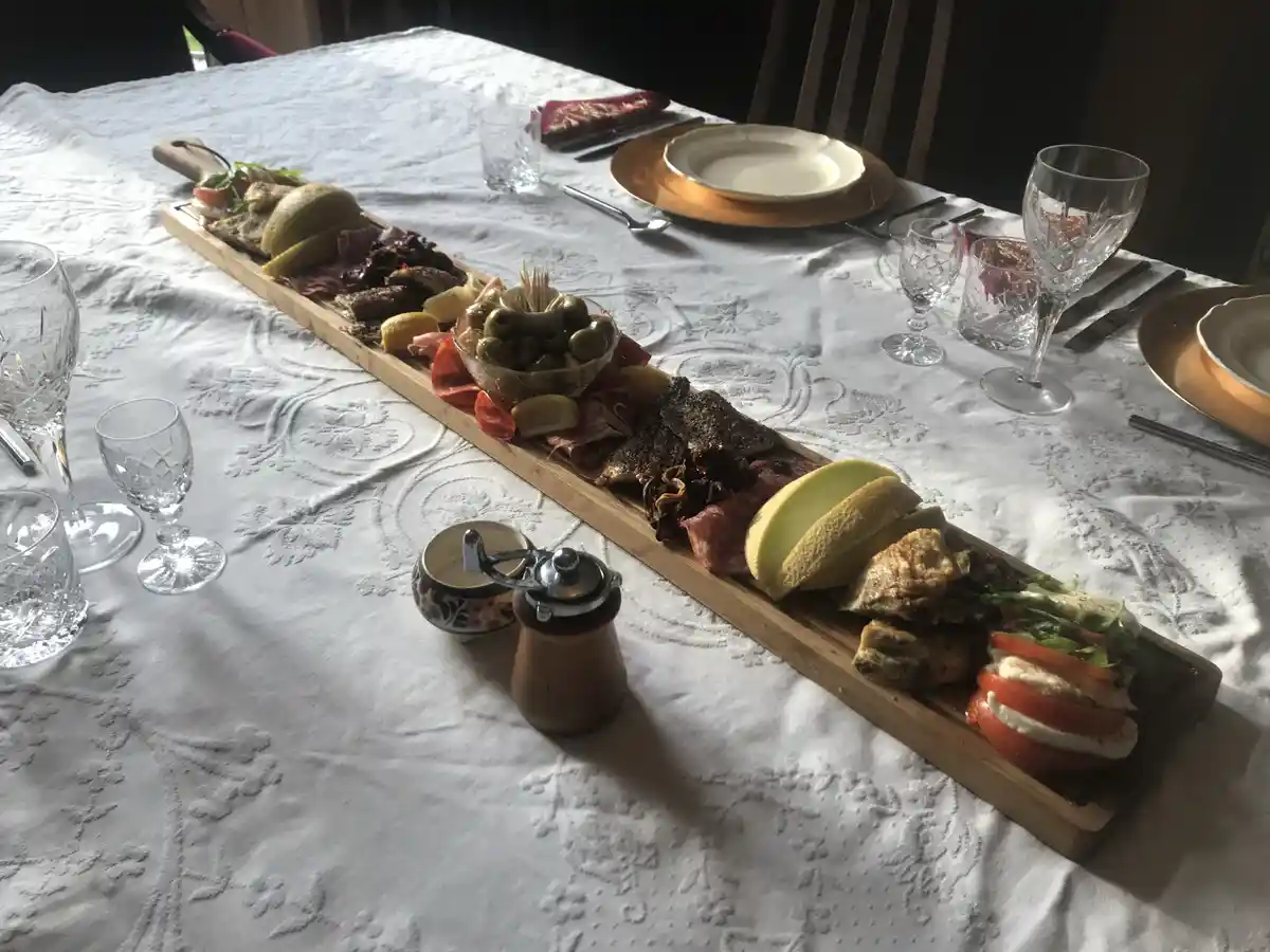 A long wooden serving board is laden with an elaborate spread of appetizers including olives, cured meats, cheese, melon…