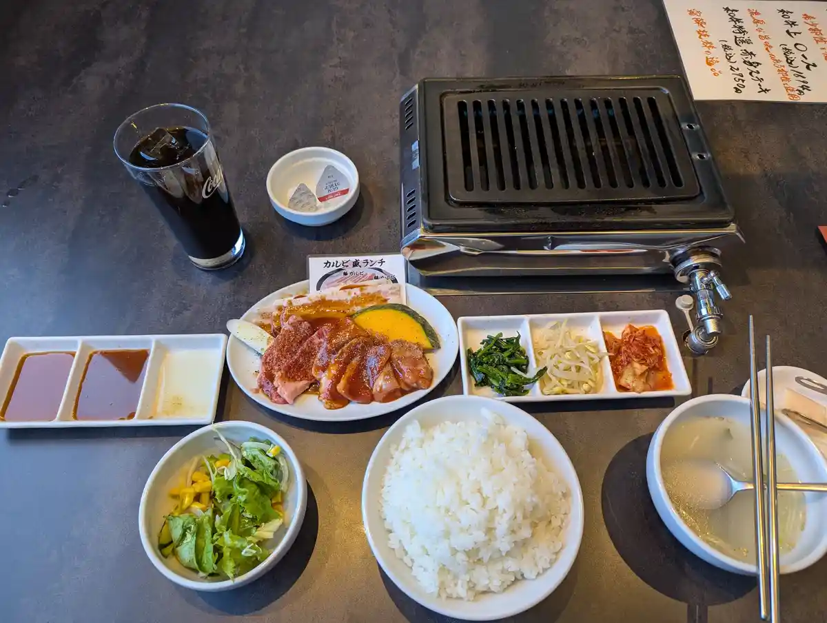 A table set with a Korean BBQ meal including a built-in grill, a plate of marinated meat, a large bowl of white rice, side…