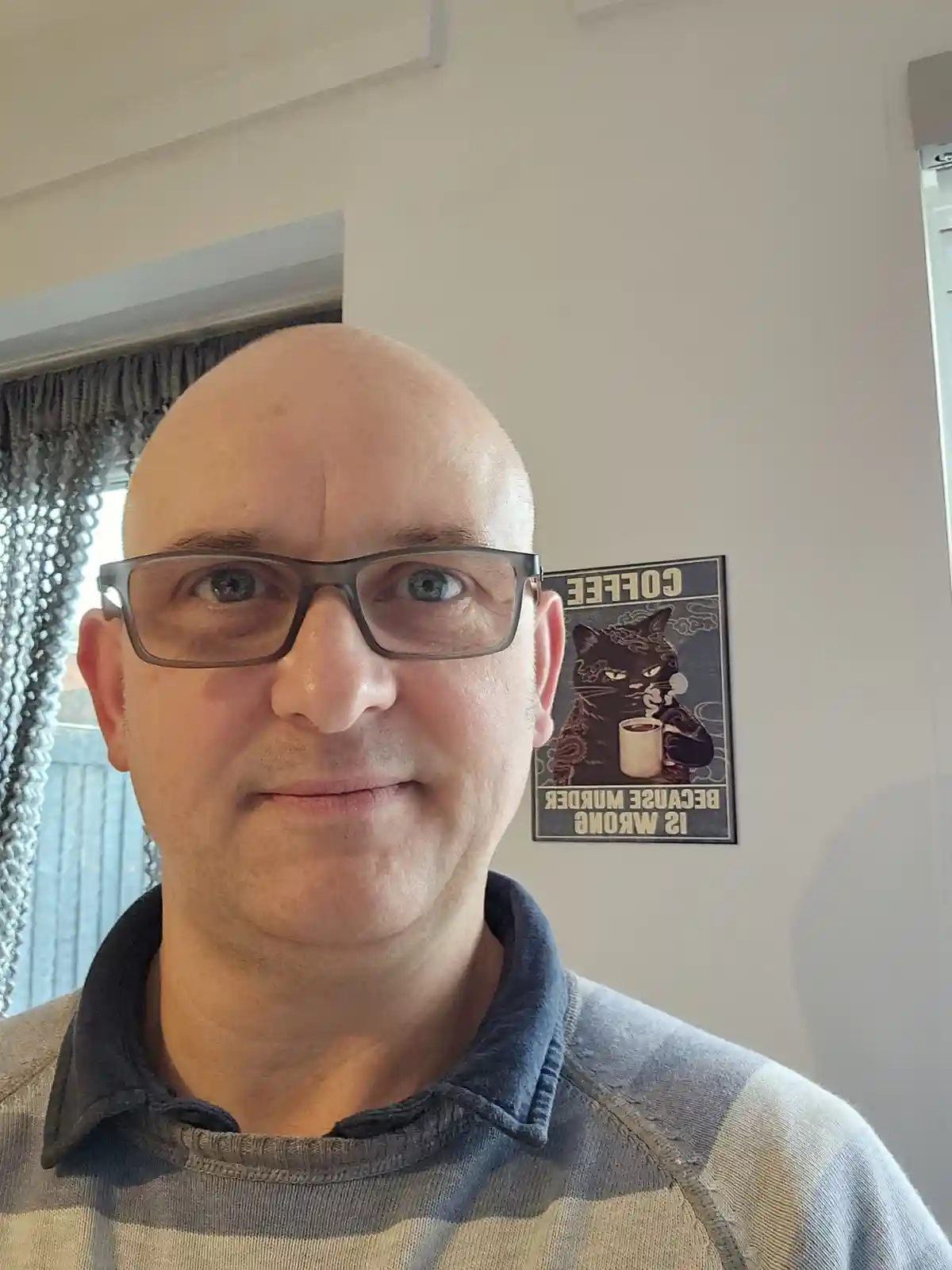A bald man with glasses stares directly at the camera with a subtle smirk, wearing a striped polo shirt. Behind him on the…