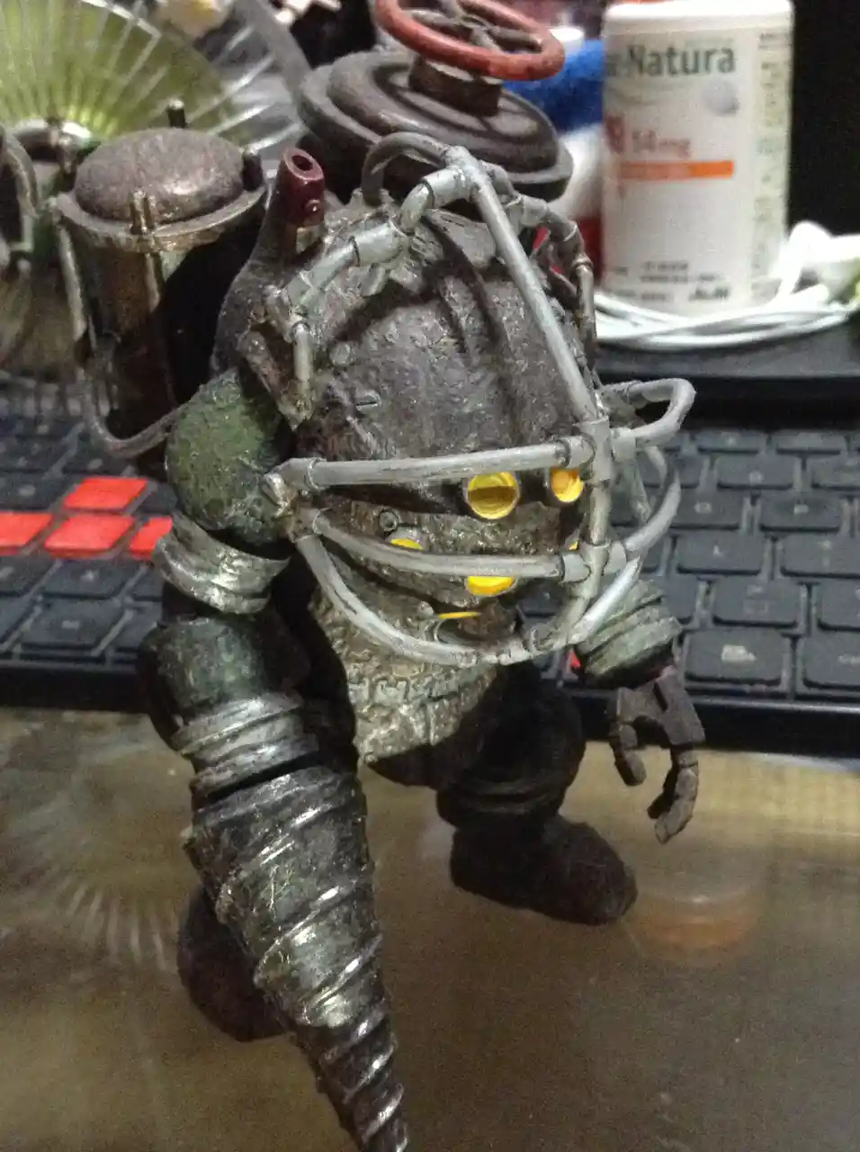 A heavily weathered, steampunk-style action figure with glowing yellow eyes and exposed pipes stands on a desk in front of a…