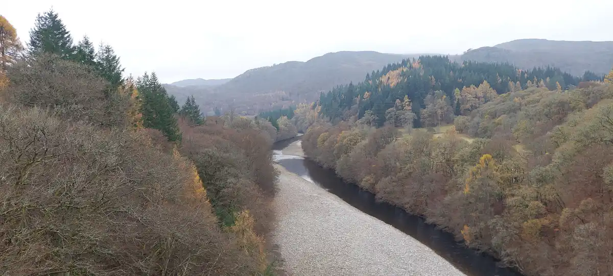 A wide-angle photo of a river winding through a forested valley in autumn, with trees showing yellow, brown, and green…