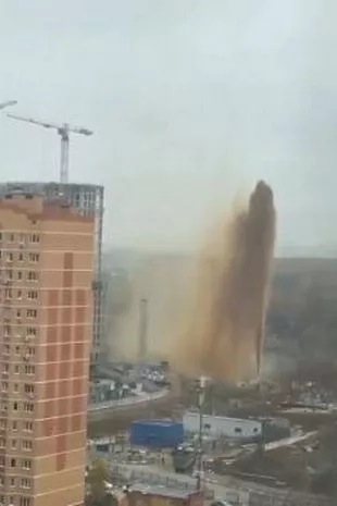 A tall, unfinished building with a crane on top is being demolished, sending a massive, towering plume of dust skyward. The…
