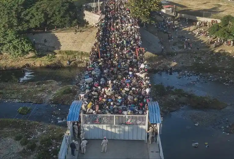 An aerial view shows a massive, dense crowd of people packed tightly across a narrow bridge over a river, many carrying…