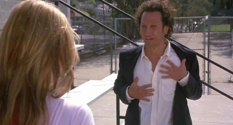 A man with curly hair and a black blazer over a white shirt gestures emphatically while speaking to a woman with blonde hair…