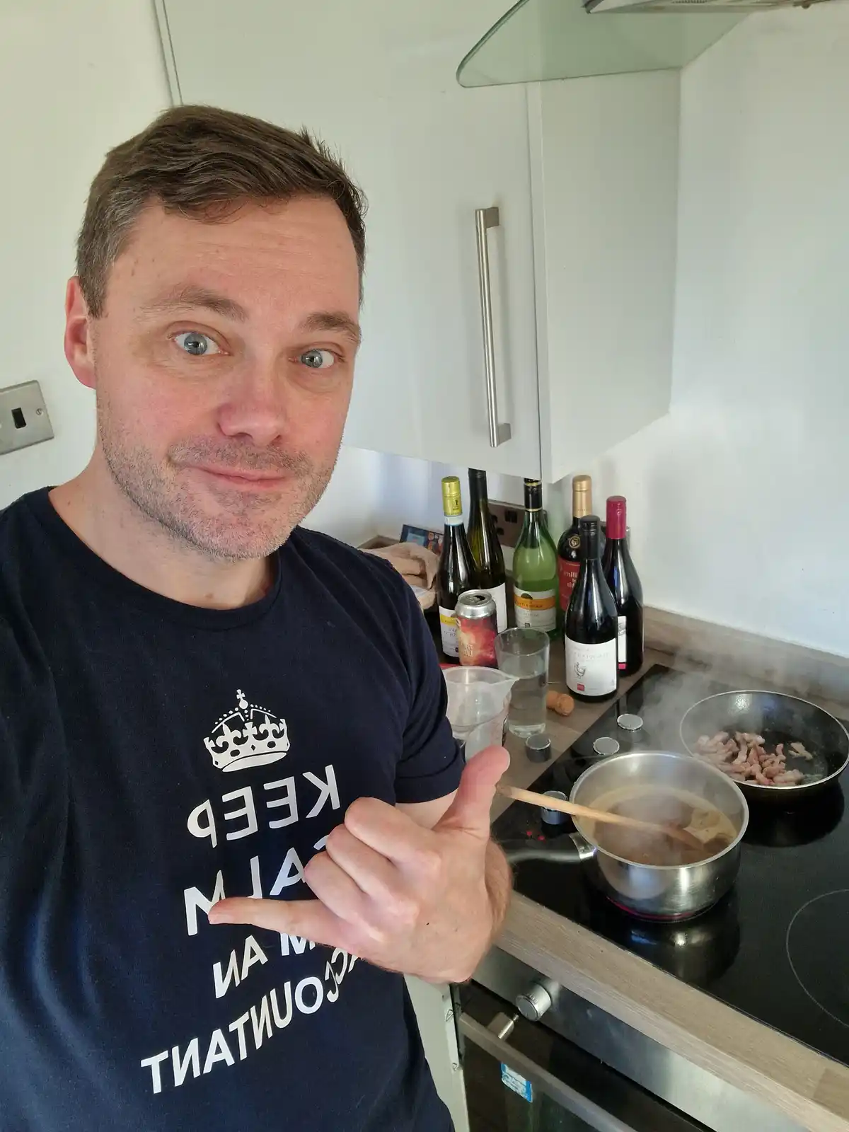 A man in a kitchen wearing a t-shirt that reads 'keep calm i am a countant' (mirrored text) gives a thumbs-up while standing…
