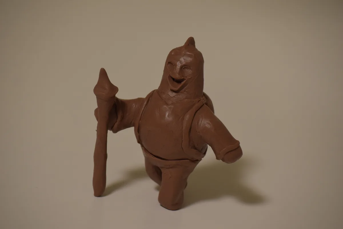 A crude brown clay figurine of a humanoid warrior with a helmet, holding a spear, and smiling broadly against a plain…