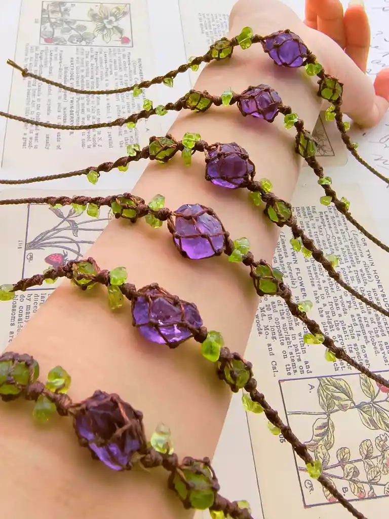 A person's wrist displays multiple handmade macrame-style bracelets featuring large purple amethyst-like stones and smaller…