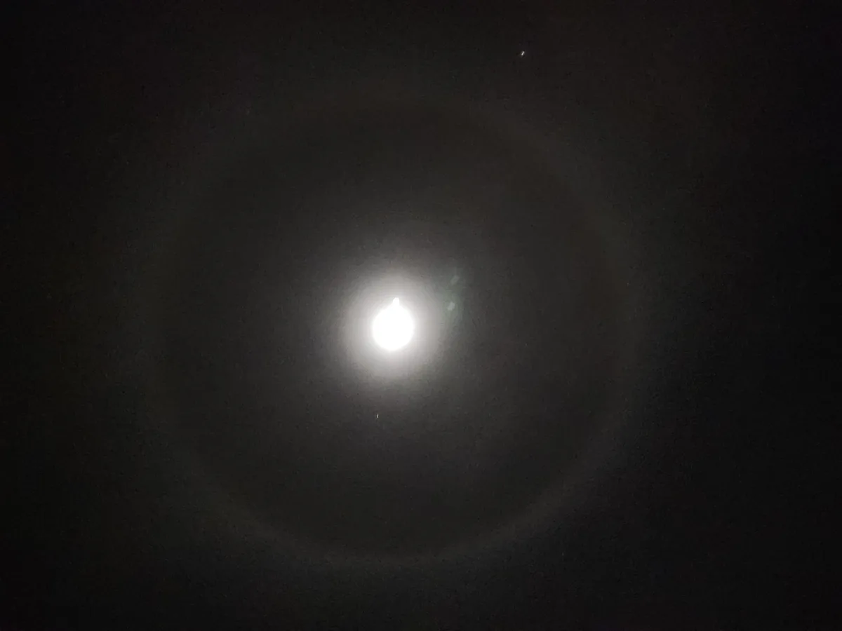 A dark night sky with a bright, slightly overexposed moon at the center, surrounded by faint atmospheric halos and a few…