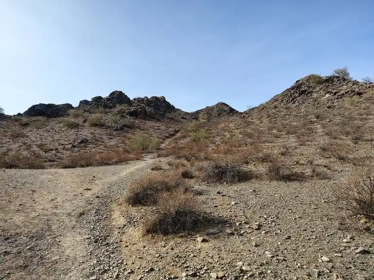 A wide-angle photo of a dry, rocky desert landscape with sparse shrubs and a dirt trail leading up to rocky hills under a…