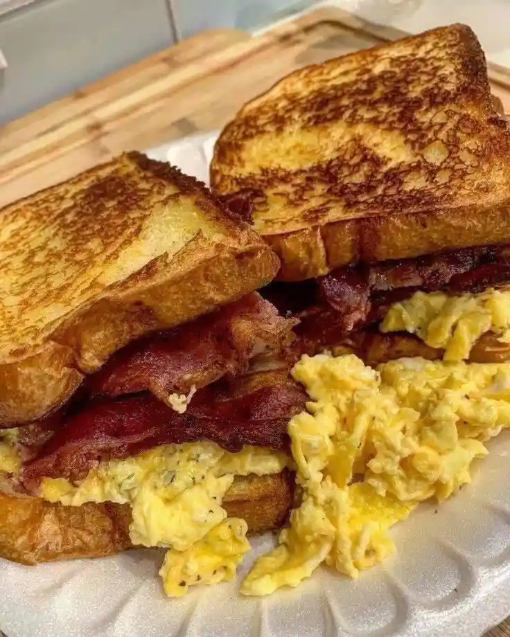 A close-up photo shows a breakfast sandwich made with two slices of golden-brown toasted bread, layered with crispy bacon…