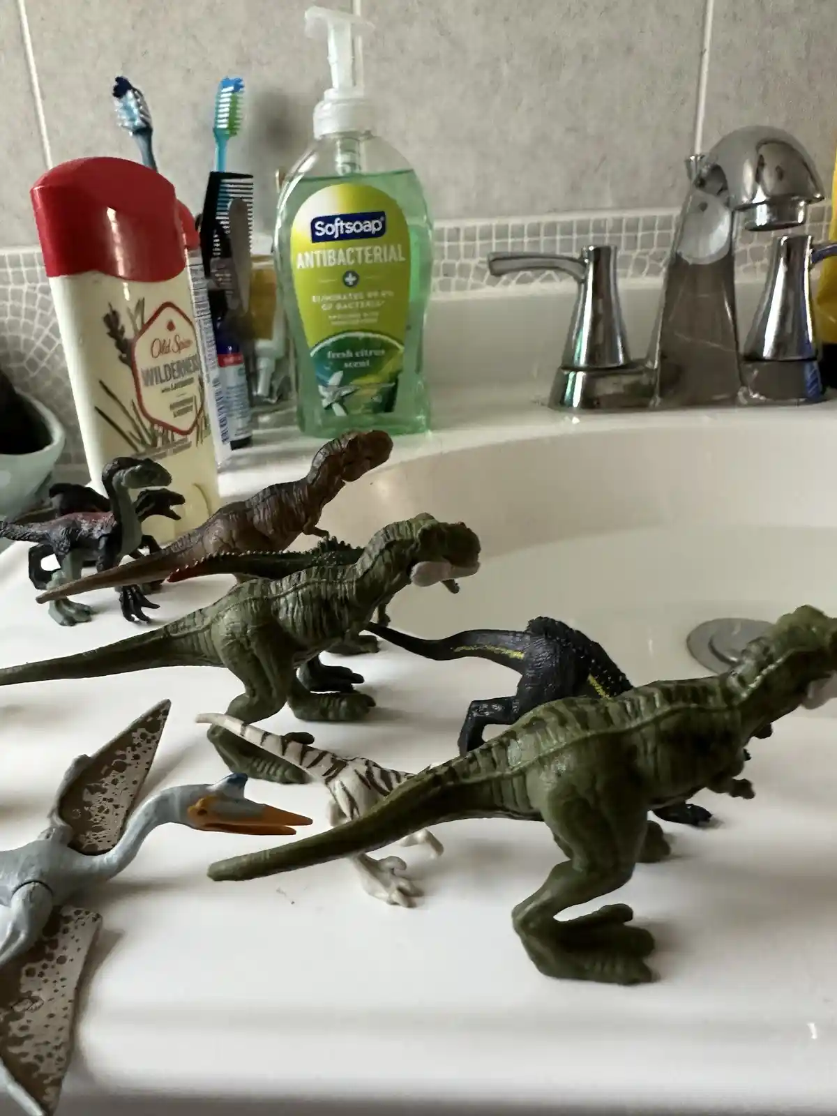 Several plastic dinosaur toys are arranged in a white bathroom sink, surrounded by everyday items like antibacterial soap…