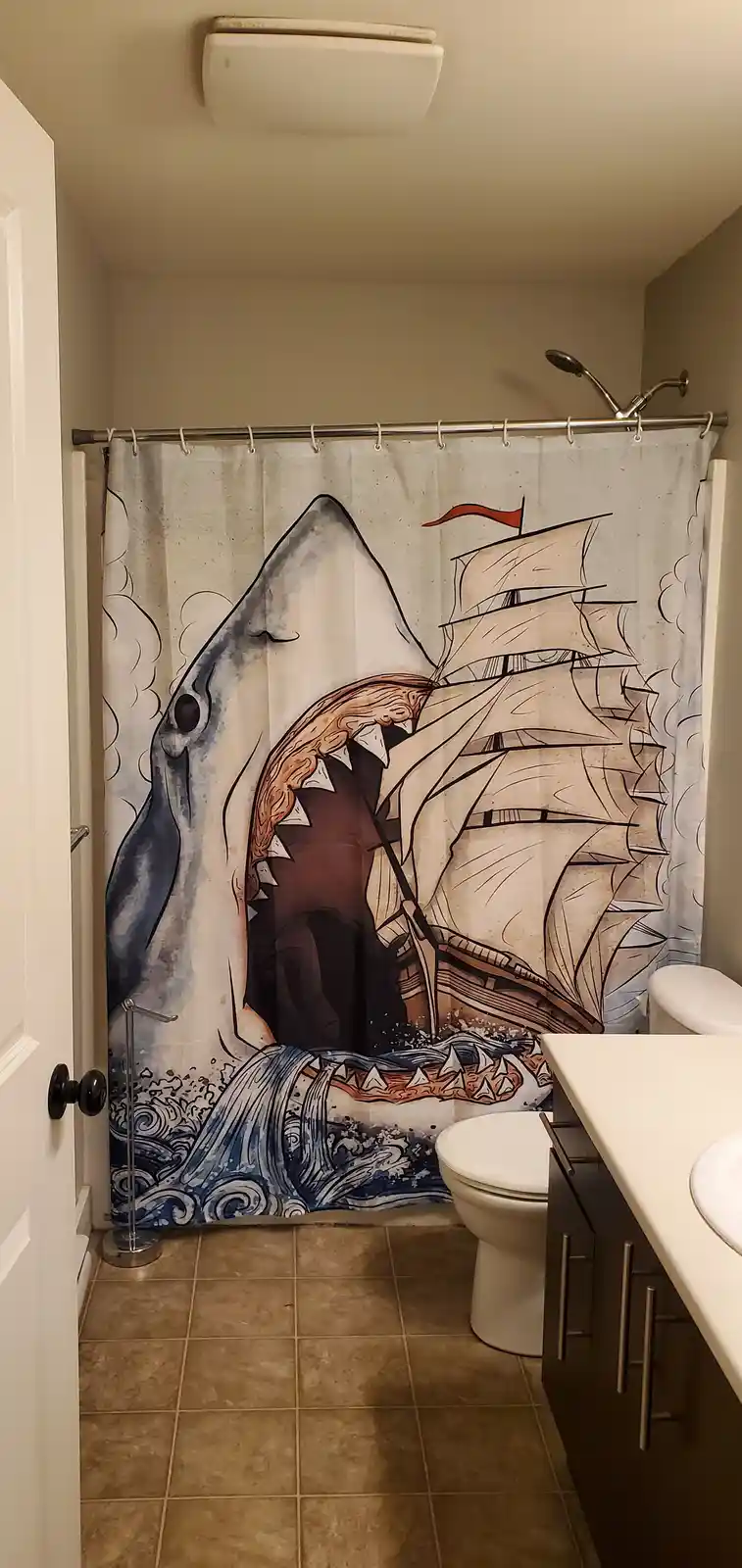 A bathroom with a shower curtain depicting a giant shark attacking a sailing ship, with a toilet and sink visible. The text…