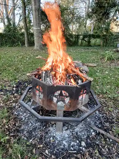 A metal fire pit with a Texas star emblem on its front is burning brightly in a grassy yard, with flames shooting upward and…