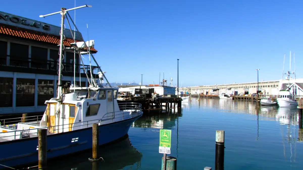A harbor scene with boats docked near a building labeled 'NOLA'S', featuring a green John Deere parking sign in the water…