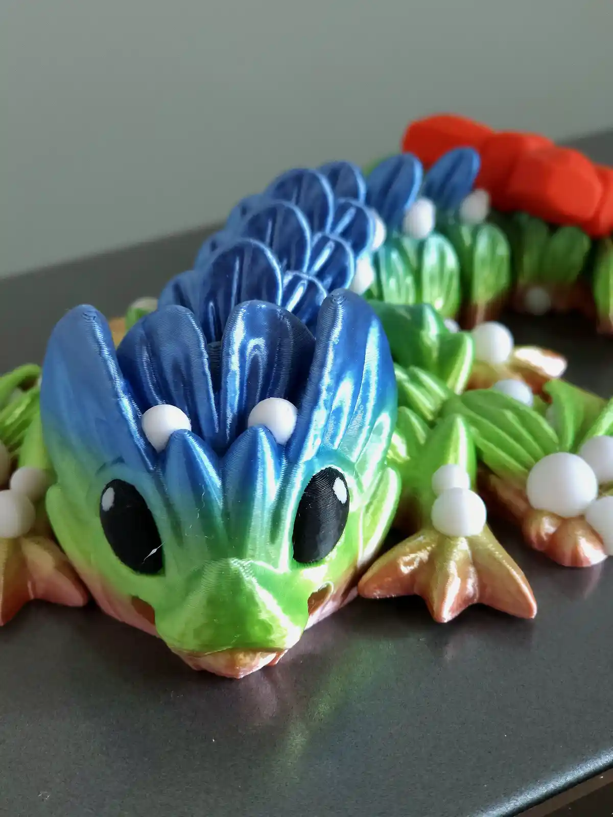 A close-up photo shows a colorful 3D-printed dragon figurine with a green head, blue spiky crest, and green body, resting on…