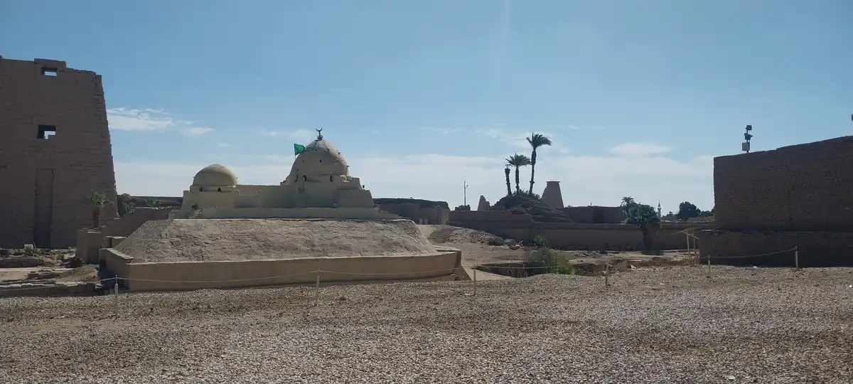 a wide-angle photo shows ancient egyptian temple ruins under a clear blue sky, with a modern white-domed mosque structure…