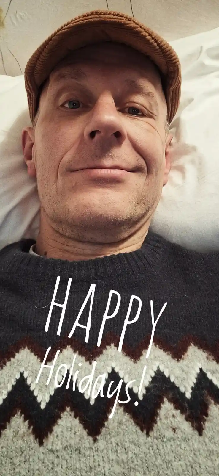 A man with a brown knit cap and a patterned sweater lies in bed, smiling at the camera. Overlaid text reads 'HAPPY…