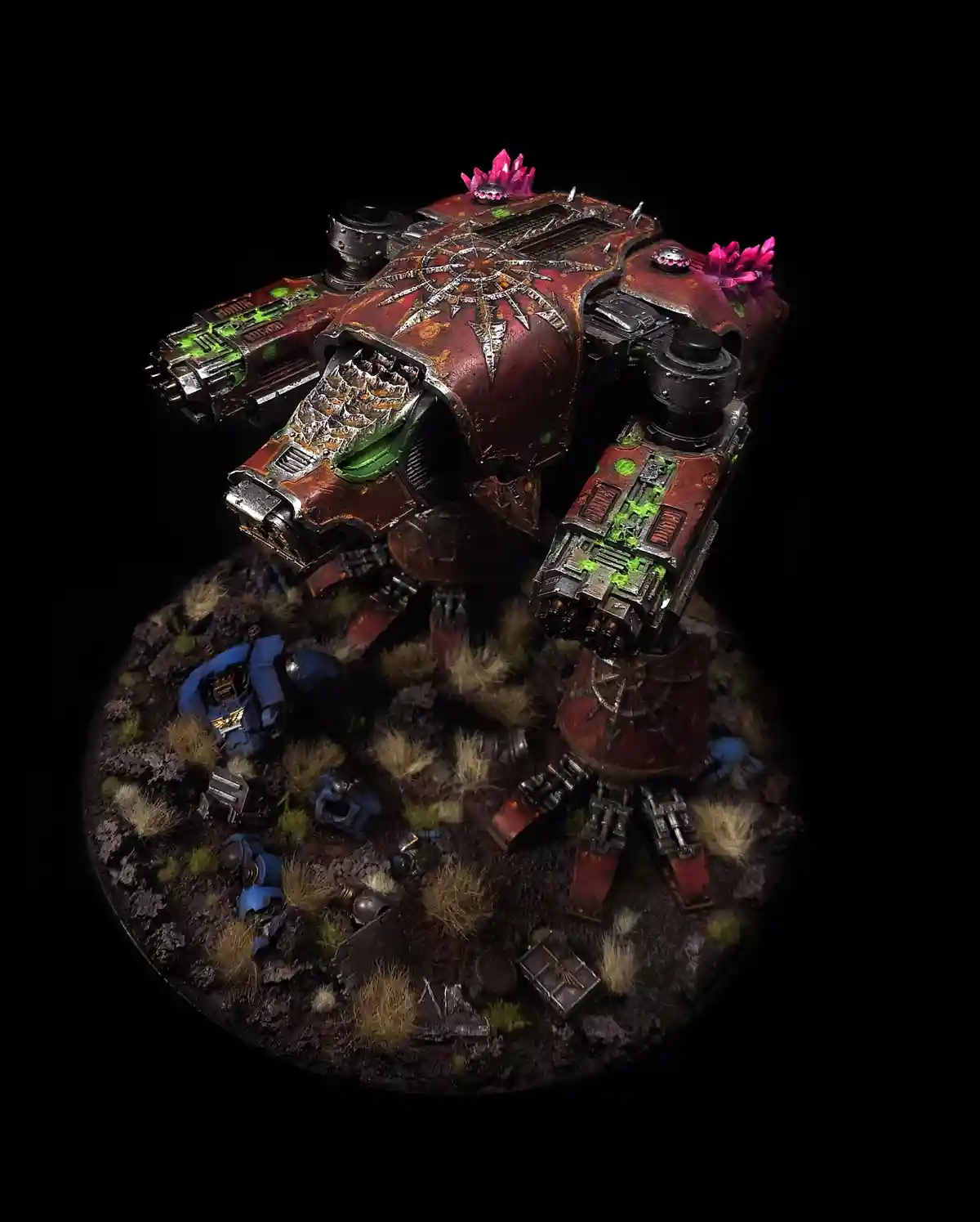 A heavily detailed, red and green painted Warhammer 40k mega-robot stands over a circular base littered with blue-armored…