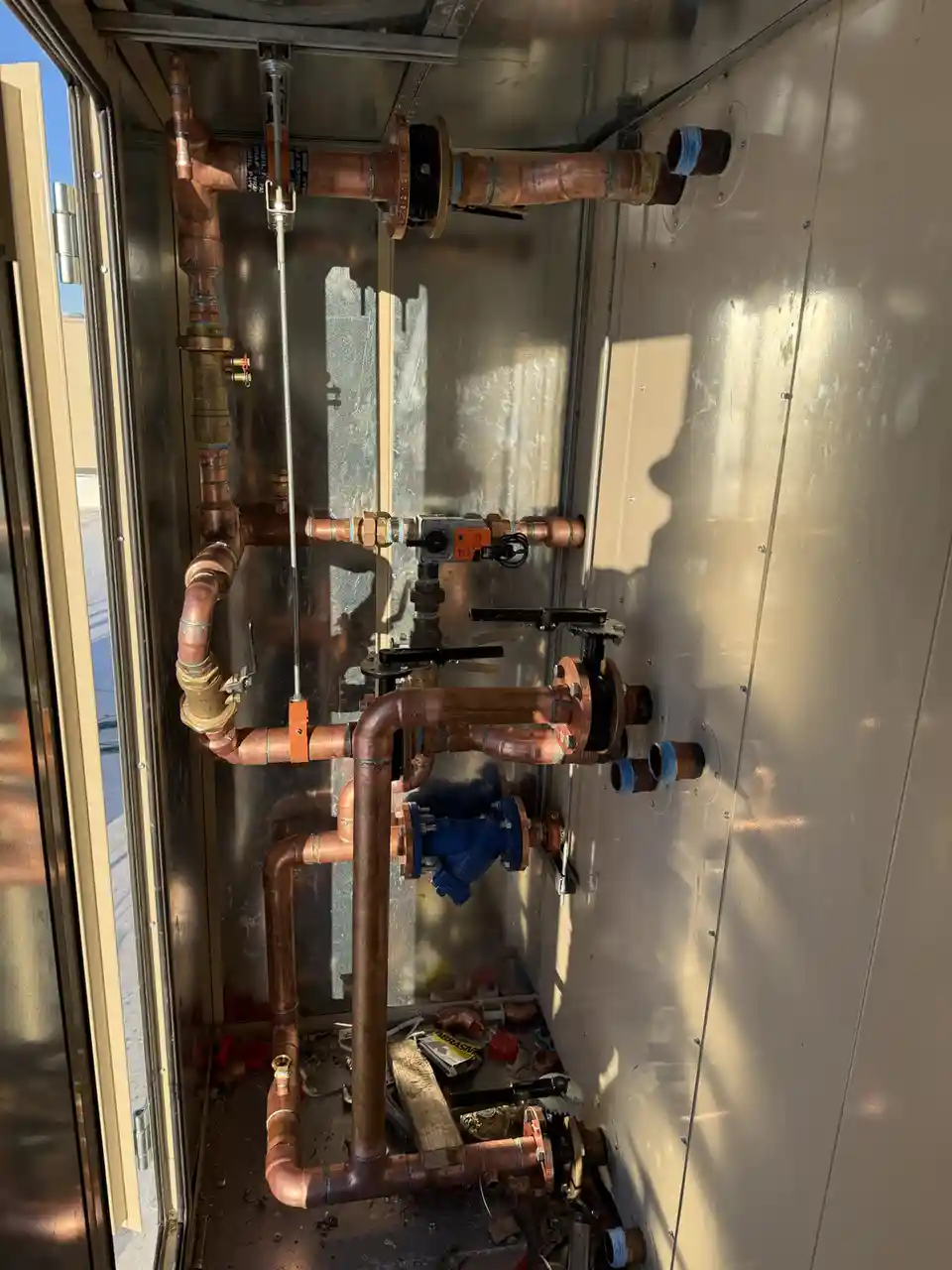 A complex network of copper pipes, valves, and fittings is installed inside a metal enclosure, with tools and debris on the…