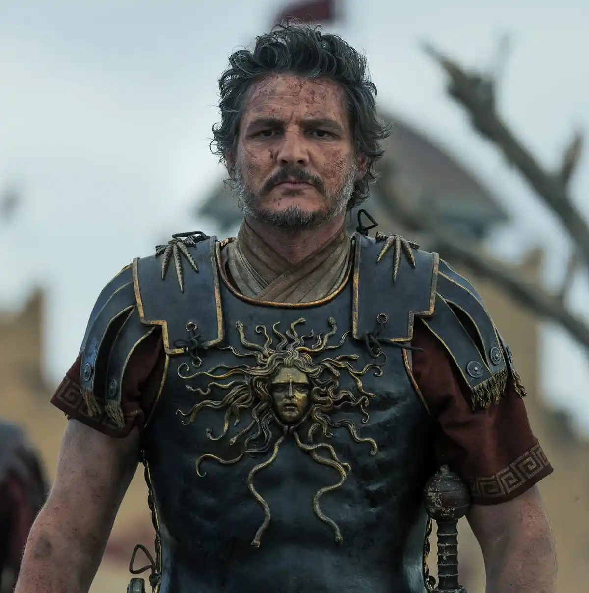 A man with a weathered, blood-streaked face and a grizzled beard wears ornate, dark armor with a golden Medusa head emblem…